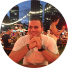 A person sits at an outdoor evening event with string lights and tall buildings in the background, smiling and resting their hands on a table, reminiscent of a professional taking a break after discussing investment loans.