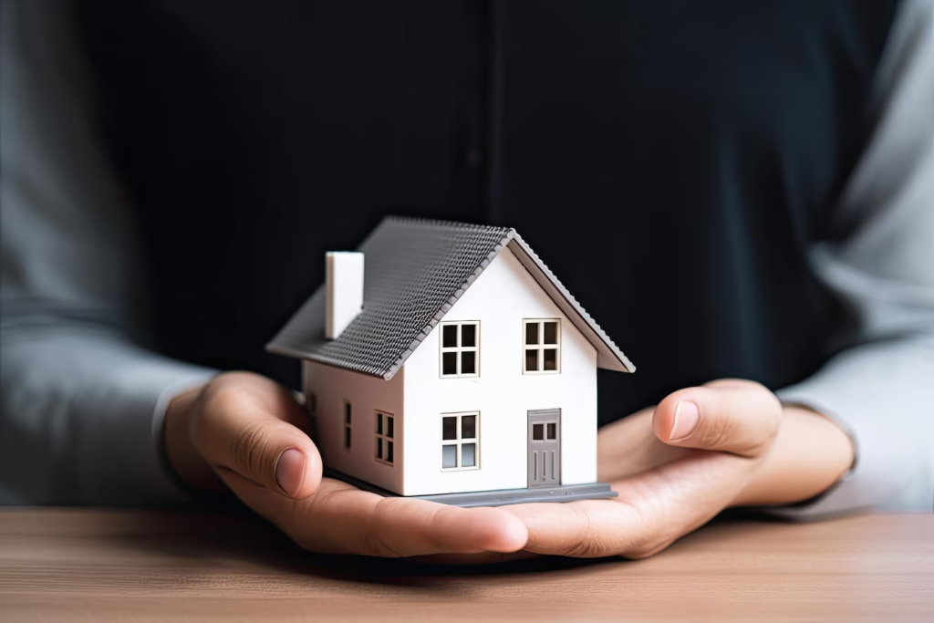 A person wearing black holds a small model house with a grey roof and white walls on a table, symbolizing obtaining a home loan for your dream property.