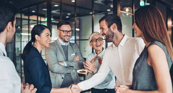 A group of professionals in business attire stand in an office, smiling and interacting. .