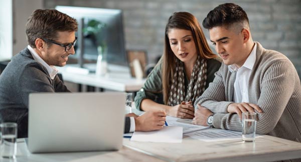 A couple sits at a table with a financial advisor, reviewing documents and discussing information about investment loans. There is a laptop and glasses of water on the table.