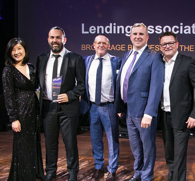 A group of five people, three men, and two women, dressed in formal attire, stand together on a stage smiling. One man holds an award plaque for excellence in Commercial Loans. The backdrop displays text that includes "Lending" and "Brokerage.