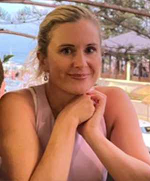 A woman with blonde hair is seated at a table with her hands clasped under her chin, looking toward the camera. She is wearing a pink sleeveless top. The background includes outdoor scenery with trees.