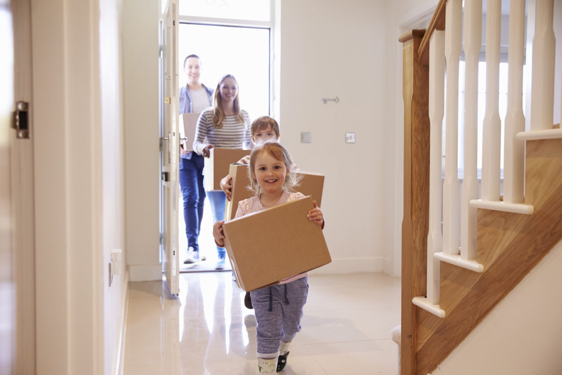 A family of four, two adults and two children, enter their new home while carrying cardboard boxes. The children lead the way, smiling.