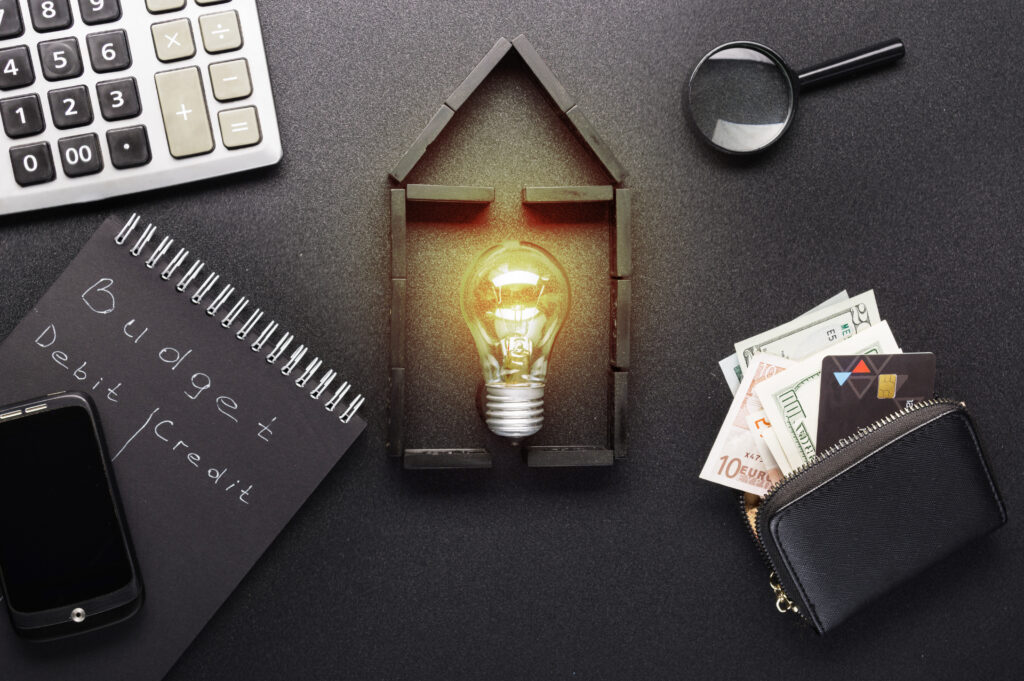 A glowing light bulb shaped like a house is surrounded by a calculator, a magnifying glass, cash, a wallet, a smartphone, and a notebook with "Budget Debit Credit" written on it—an illustration symbolizing the essentials of managing home loans.