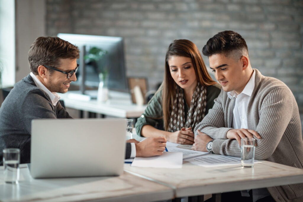 Three people sit at a table in an office, looking at paperwork together. Two of them are across from the person with a laptop.