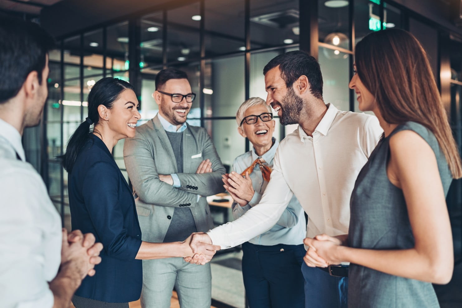 Five people in business attire are smiling and shaking hands in a modern office setting with glass walls.