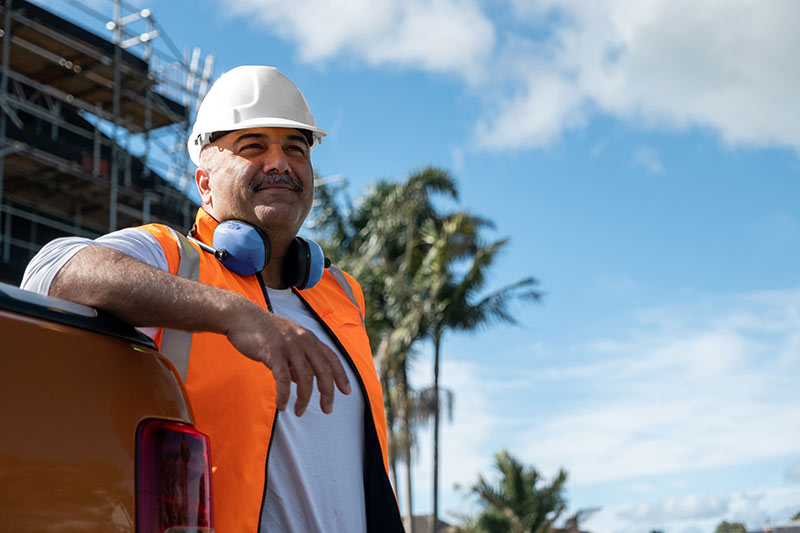 A construction worker in an orange safety vest and white hard hat stands next to a vehicle, with a building site and palm trees in the background.