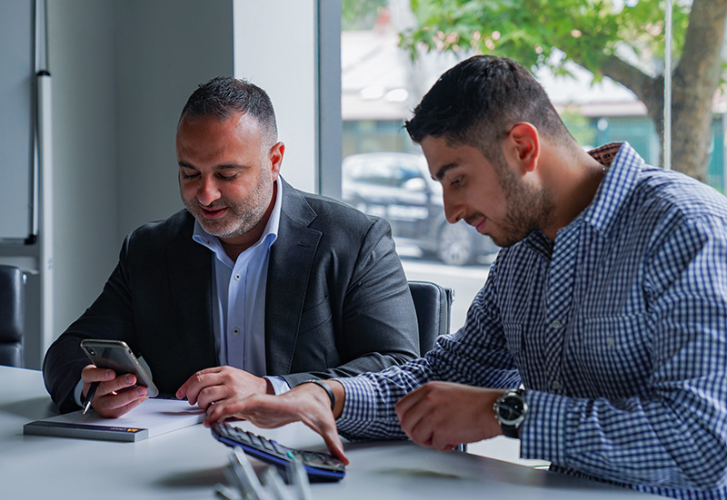 Two men are seated at a table in an office setting. One is looking at his phone and the other is writing on a notepad. There is a window with a view of trees in the background.