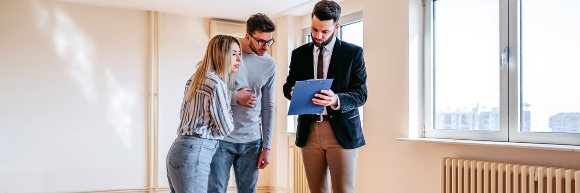 A man in a suit shows a clipboard to a man and woman in a brightly lit room with large windows and radiator heaters, discussing options for home loans.