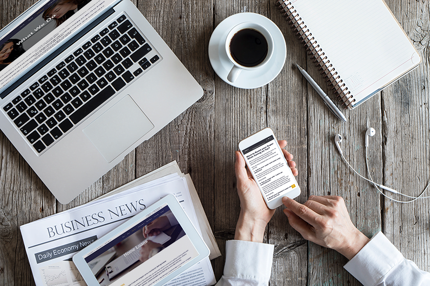 A person reading news on a smartphone with a laptop, tablet, coffee cup, and earphones on a wooden table while researching Home Loans.