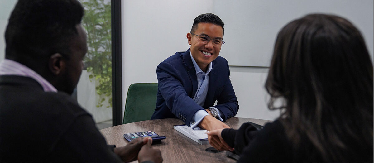 Three people are seated at a table in a business meeting. The man in the center, wearing a suit, is smiling and shaking hands with a person on the right.