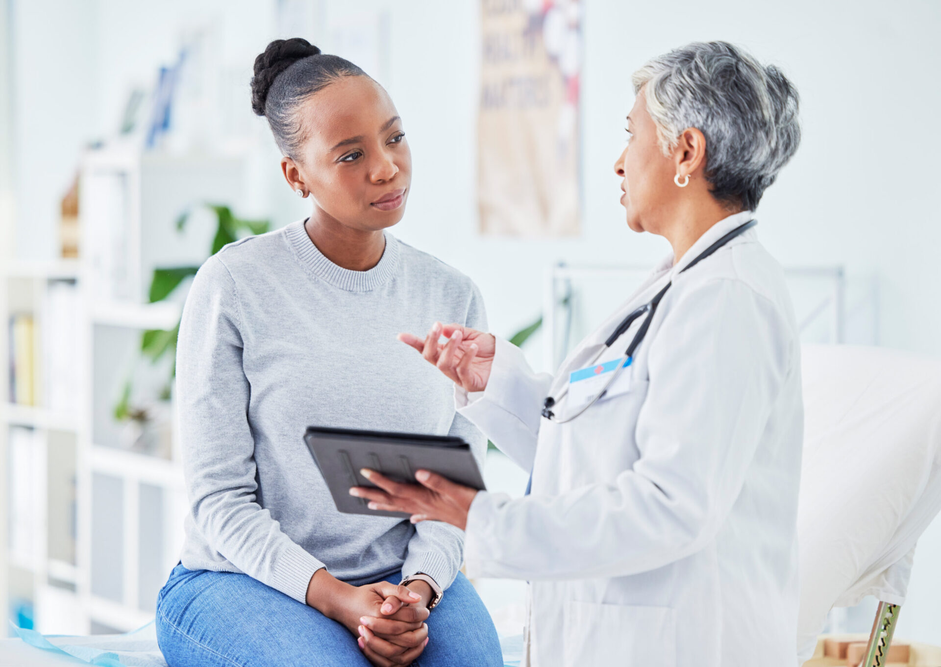 A doctor in a white coat talks to a patient who is sitting on an examination table, holding a tablet. The patient listens attentively, as if discussing the complexities of investment loans.