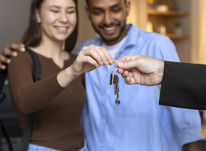 A person hands over a set of keys to a smiling couple standing closely together, their joy reflecting the culmination of securing their dream through home loans.