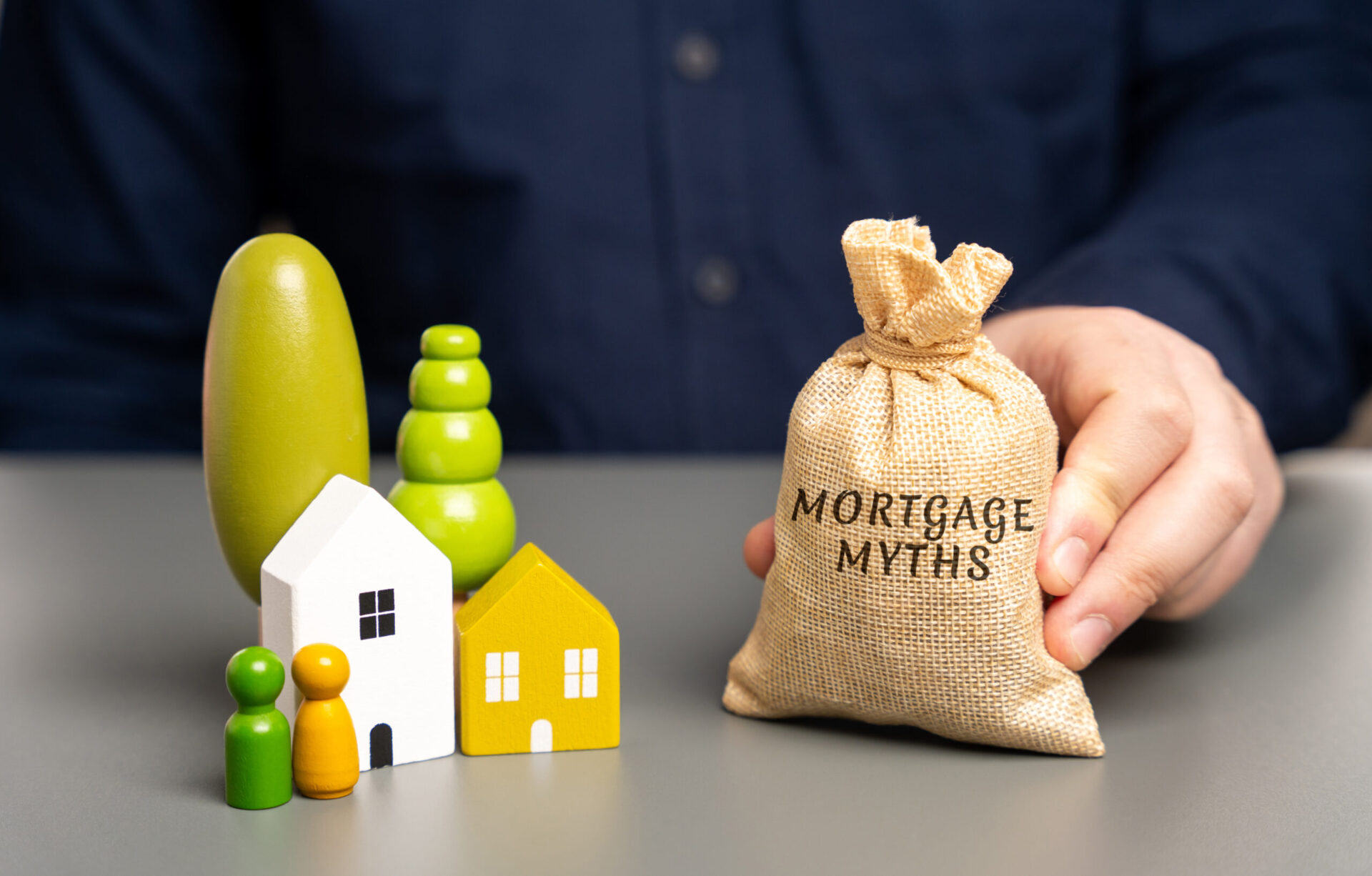 A person holding a burlap sack labeled "Mortgage Myths" stands next to small wooden figures of houses, trees, and people on a table