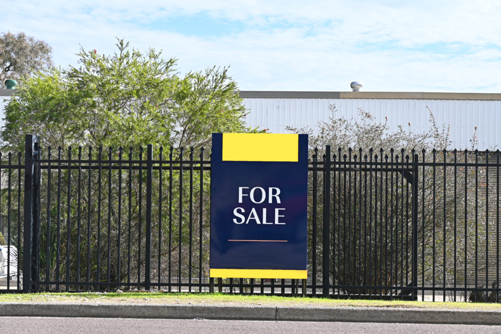 A large "For Sale" sign is attached to a black metal fence, with trees and a building visible in the background.