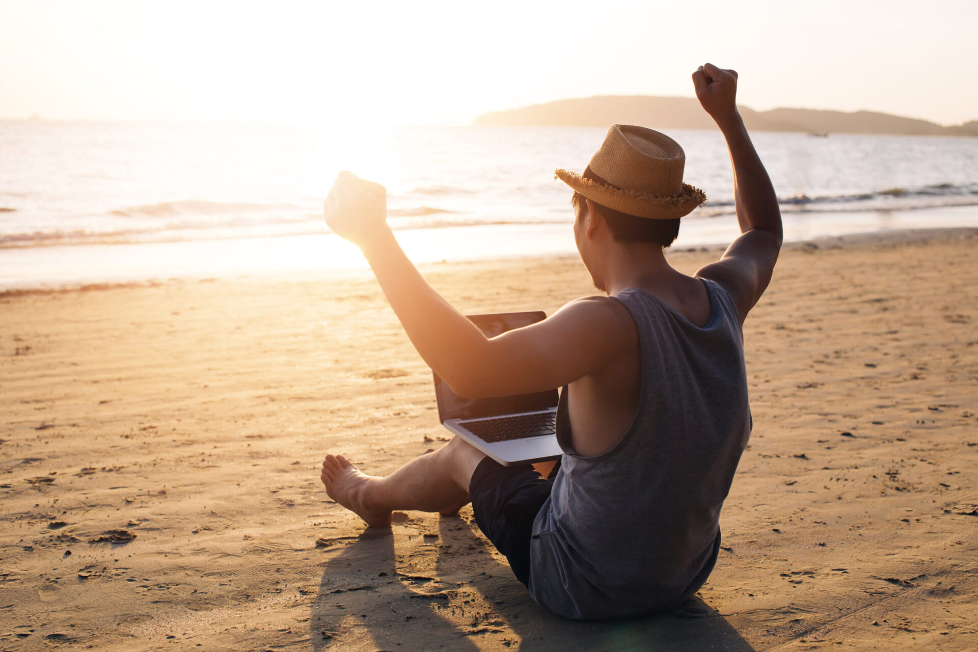 A person wearing a hat and a sleeveless shirt sits on a beach with a laptop, raising their arms in a celebratory gesture as the sun sets over the ocean, perhaps having just secured favorable terms for construction loans.