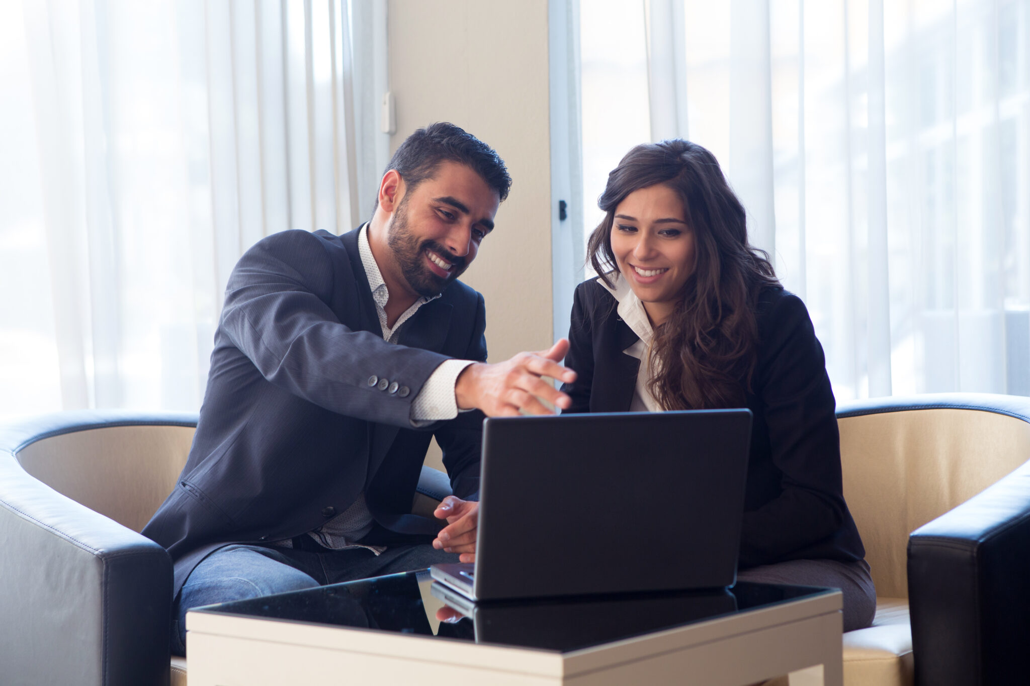 Two individuals in business attire sit in an office, smiling and looking at a laptop screen on a glass-topped table. One person is pointing at the screen, discussing options for commercial loans.