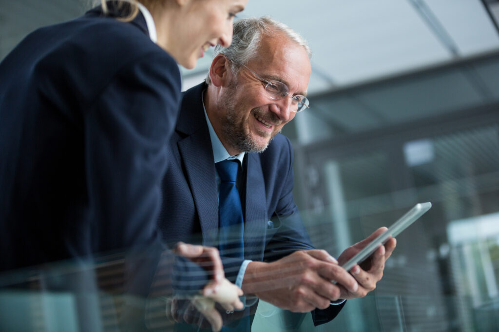 Two professionals in formal attire are smiling and looking at a tablet. They appear to be having a pleasant conversation in a modern office setting.