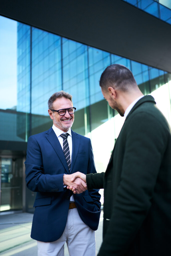 Two men in business attire shake hands outside a modern glass building.