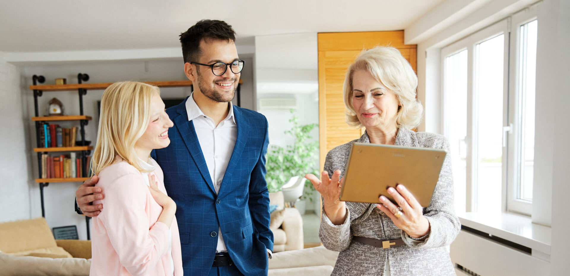 A real estate agent with a tablet shows a property to a smiling couple in a modern, well-lit living room.