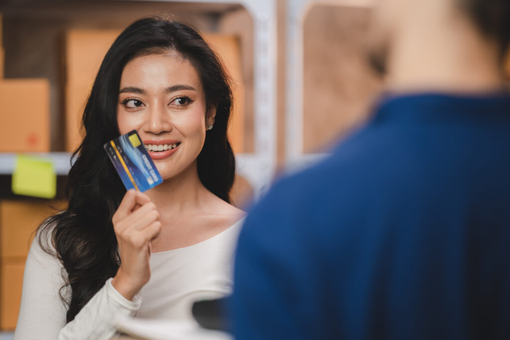 A woman holding a credit card and smiling while interacting with a person in the blurred background of a store, likely discussing commercial loans.