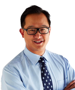 A person wearing glasses, a light blue shirt, and a dark blue polka dot tie, standing against a plain white background.