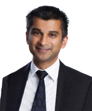 A person with short dark hair and a dark suit jacket over a white shirt and dark tie smiles against a white background.