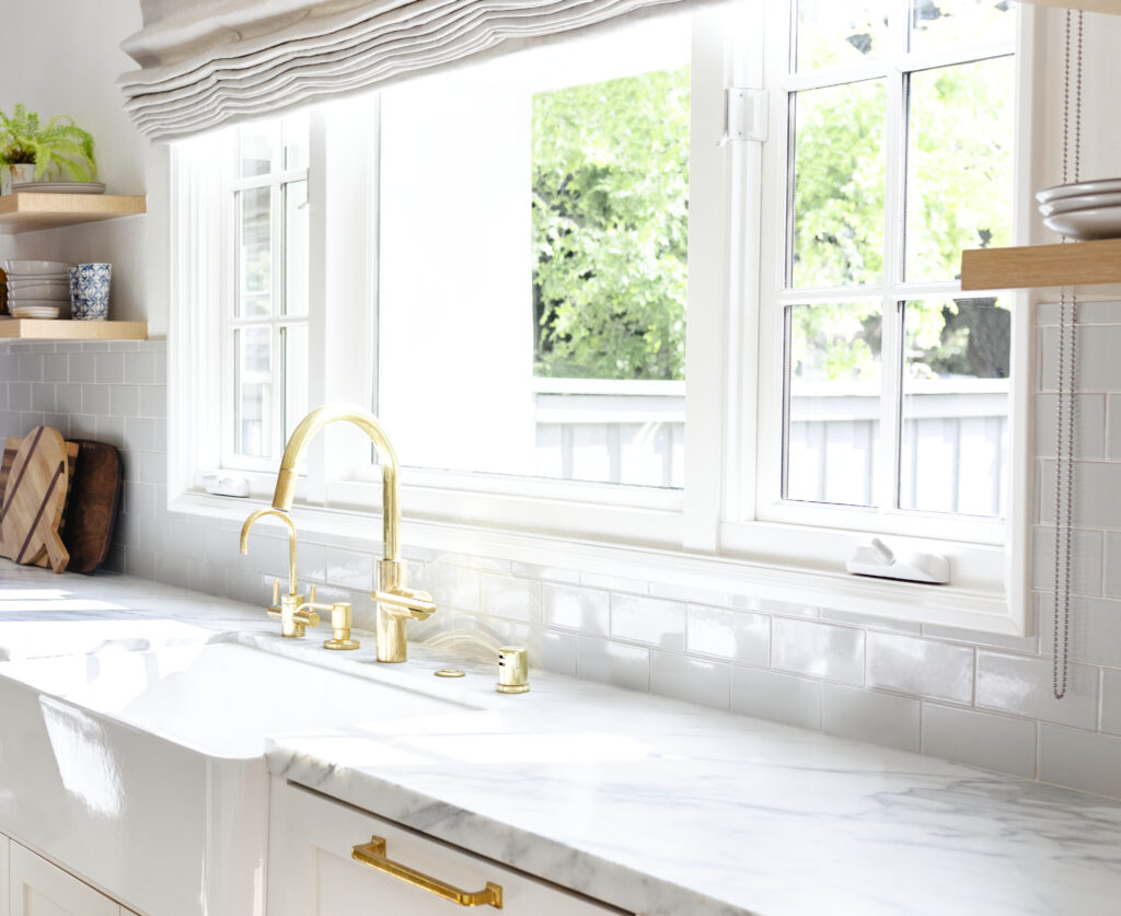 A bright kitchen, perfect for envisioning your next culinary adventure, features a marble countertop, gold faucet, white tile backsplash, and large windows with natural light streaming in—a dream made possible through versatile construction loans.