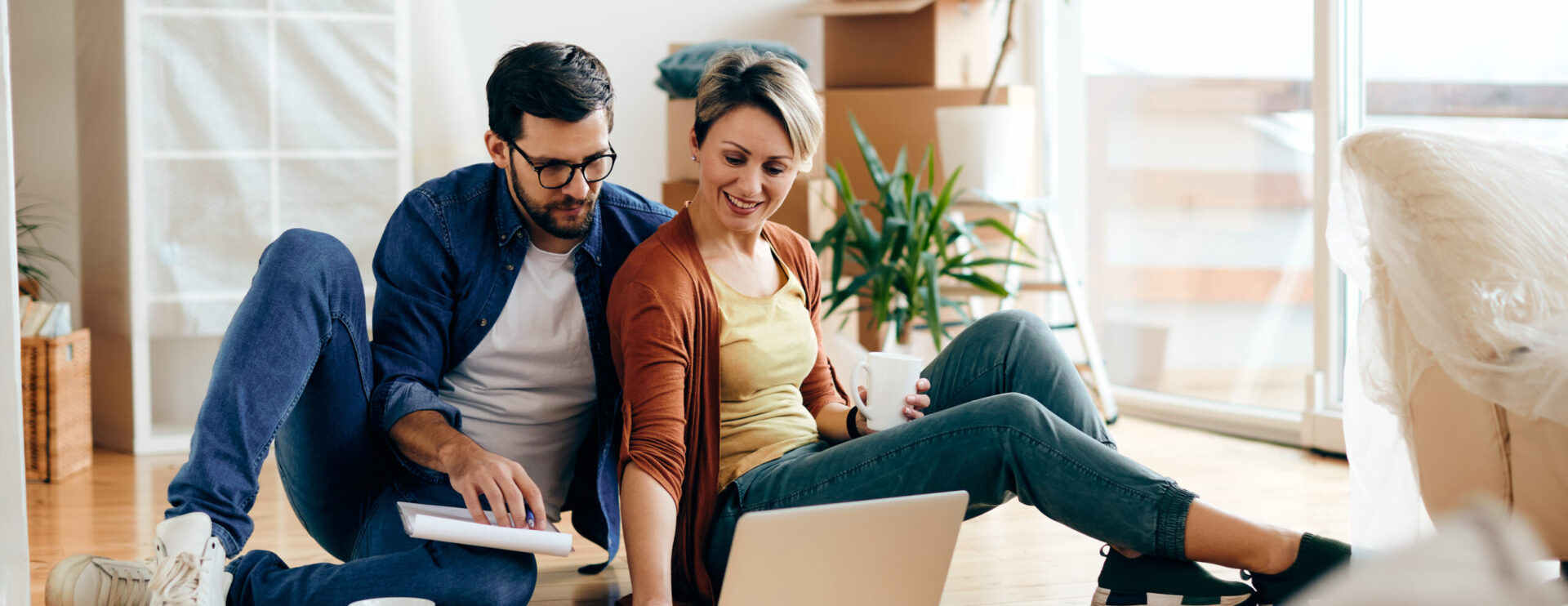 A man and a woman sit on the floor, looking at a laptop. The man holds a tablet, and the woman holds a coffee cup. Boxes and plants are visible in the background.