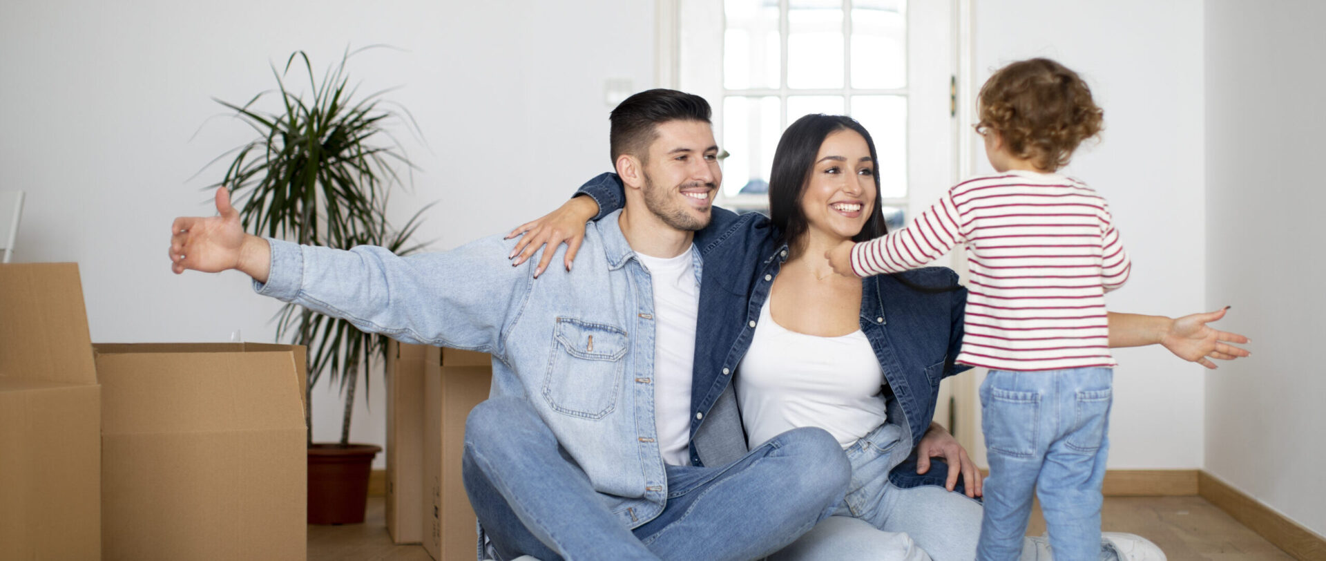 A man and a woman sit on the floor with open arms, while a child in a striped shirt walks towards them. Several unpacked boxes are in the background, symbolizing the new beginnings made possible by their home loan.