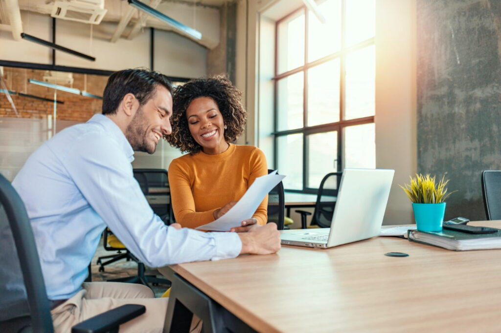 Two people sitting at a table in an office, reviewing documents and smiling; a laptop, plant, and other office supplies can be seen in the background.