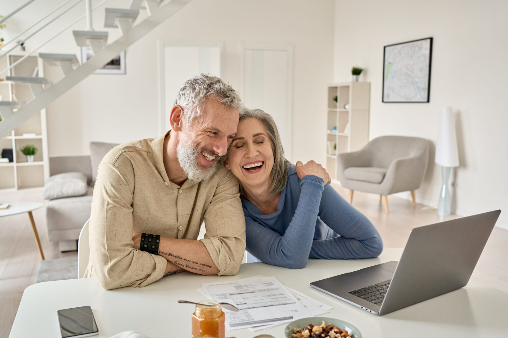 An older couple sits at a table with documents and a laptop, smiling and leaning in towards each other in a bright, modern living room, possibly discussing options with Lending Association.