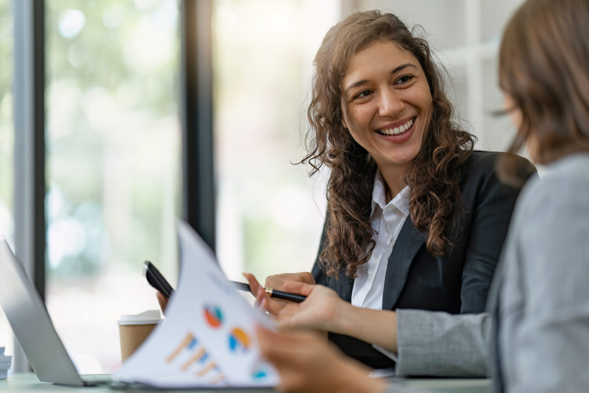 Two people in business attire are sitting at a desk, smiling and talking. One person is holding a pen, while a document with charts related to Business Loans is visible in the foreground.