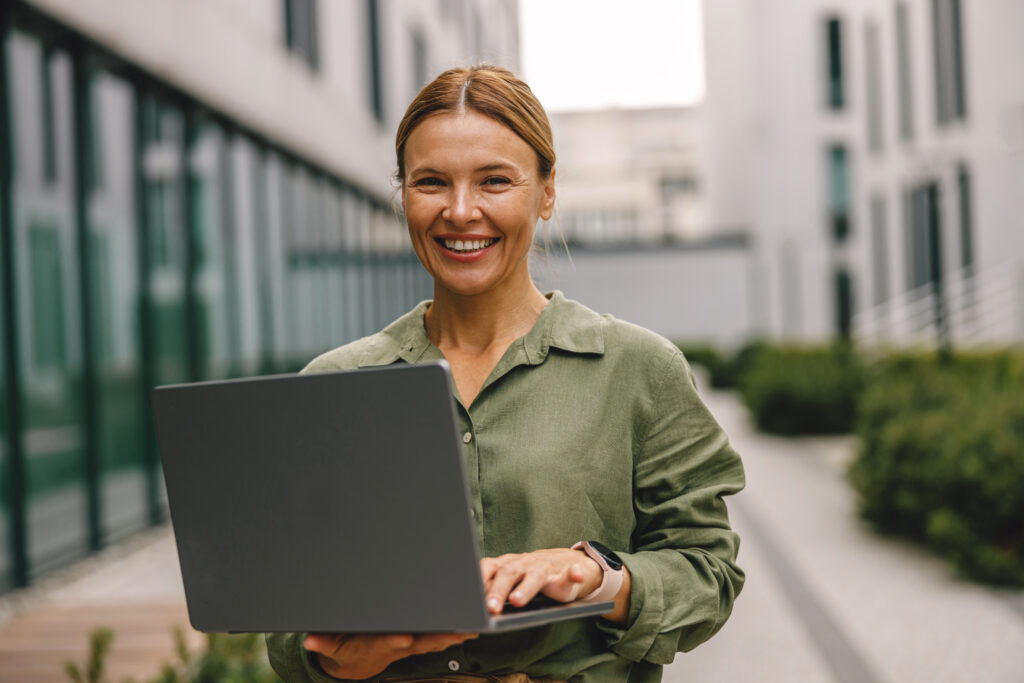 A woman stands outdoors, smiling and holding an open laptop. She is wearing a green shirt and appears to be in a modern urban setting with buildings and greenery in the background, perhaps discussing business loans online.