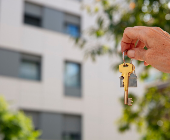 A hand holding a keyring with several keys in front of a blurred exterior of a building.