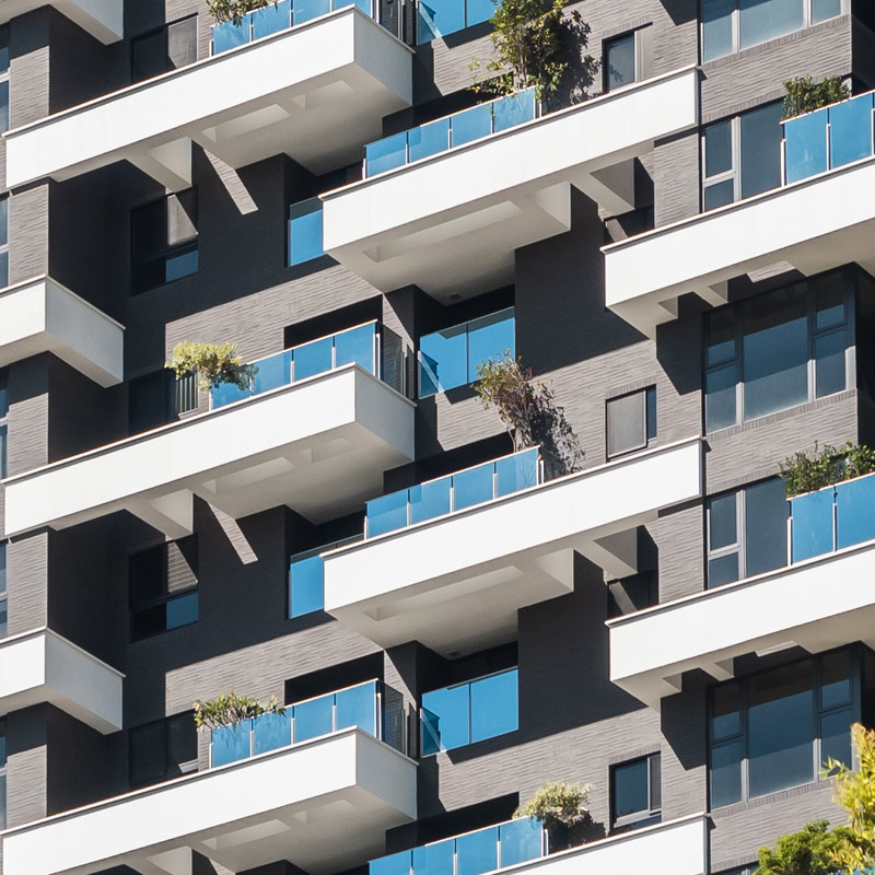 A modern apartment building with multiple balconies featuring glass railings and plants. The building has a sleek, geometric design with a mix of gray and white exterior finishes.