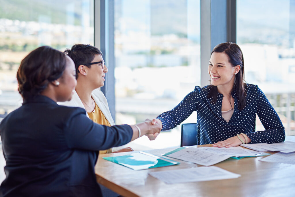 Three people having a meeting in a bright office, with one woman shaking hands with another across a table covered in business loans documents.