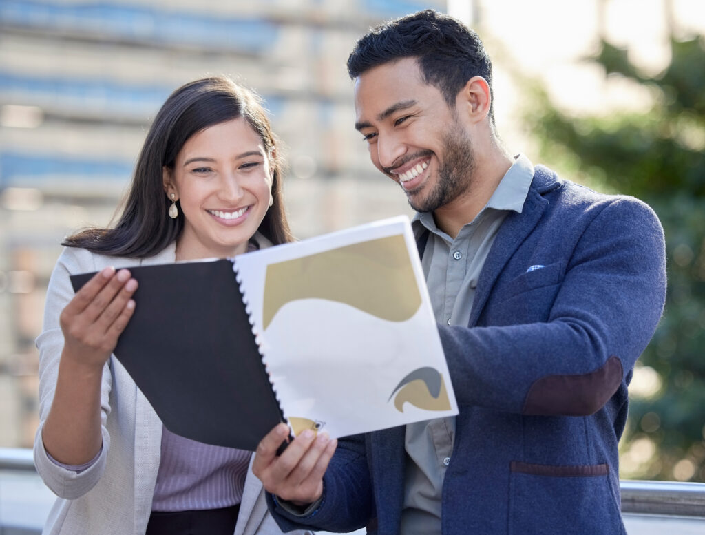 Two people smiling and looking at a document outdoors.