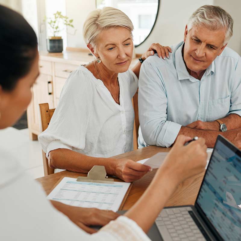 Three people at a meeting. An older couple listens attentively to a person from Lending Association showing information on a laptop screen. Documents and a form are on the table.