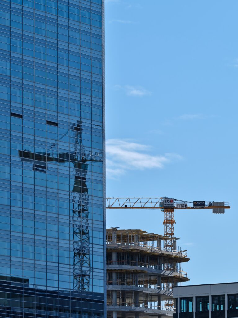 A construction crane and partially built structure are reflected in the glass facade of an adjacent tall, modern building under a clear blue sky.