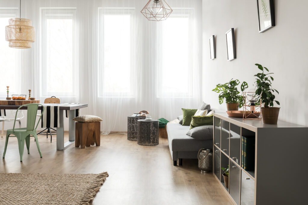 A modern living room with a gray sofa, potted plants, shelving unit, dining table with chairs, and large windows with sheer curtains. Geometric pendant lights hang from the ceiling. Considering Home Loans? This stylish space could be yours to enjoy every day.