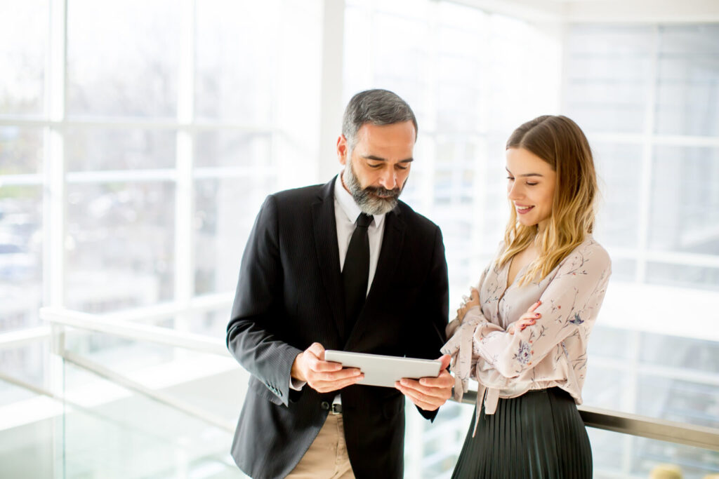 A man in a suit and a woman in a blouse stand in a modern office, discussing business loans while looking at a tablet together.