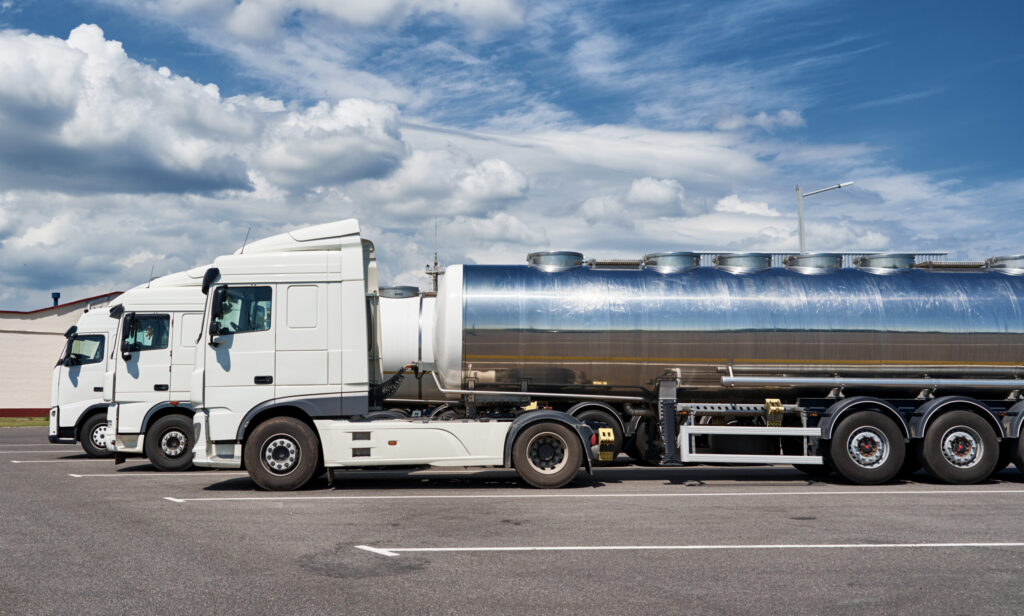 A white tanker truck with a shiny, metallic tank is parked in an open lot under a partly cloudy sky. Another similar truck is seen parked beside it.