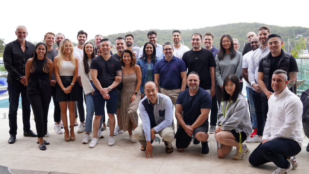 A group of 27 people standing and kneeling outdoors, posing for a photo near a pool with hills in the background. Dressed casually and some smiling, they seem ready to celebrate their success in investment loans.