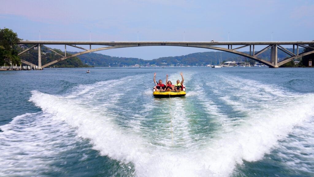 A group of people ride an inflatable tube being towed by a boat on a river, with a large bridge in the background, discussing exciting opportunities like investment loans for their future.