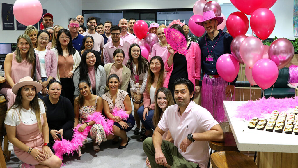 A group of people from Lending Association dressed in various shades of pink celebrate in an office adorned with pink balloons, feather boas, and cupcakes. Some individuals are seated, while others are standing close together.