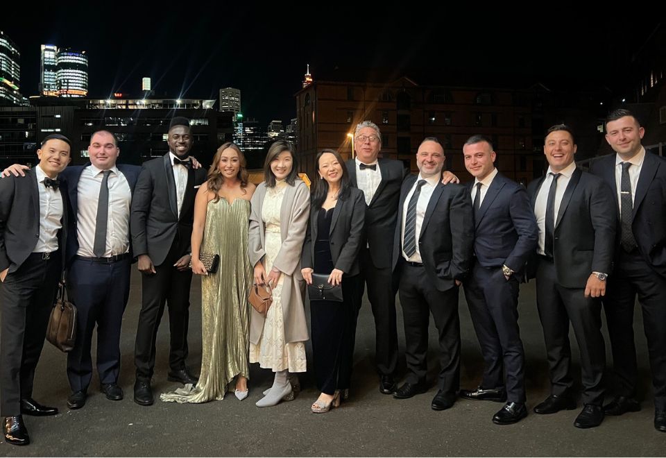 A group of 11 people dressed in formal attire standing together outdoors at night, with a cityscape in the background, possibly discussing Investment Loans.