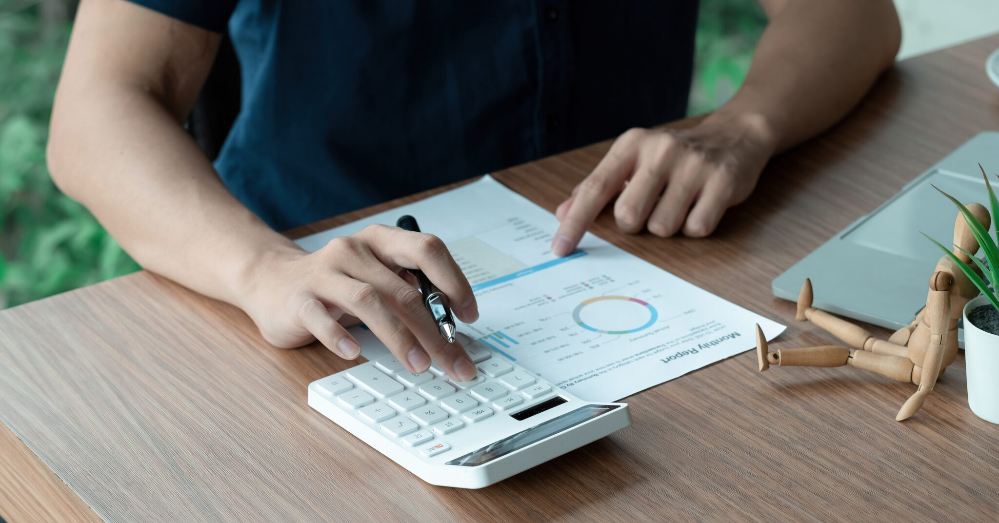 Person using a calculator and pointing to a chart on a paper on a wooden desk, next to a laptop and small plant, perhaps assessing commercial loans or construction loans