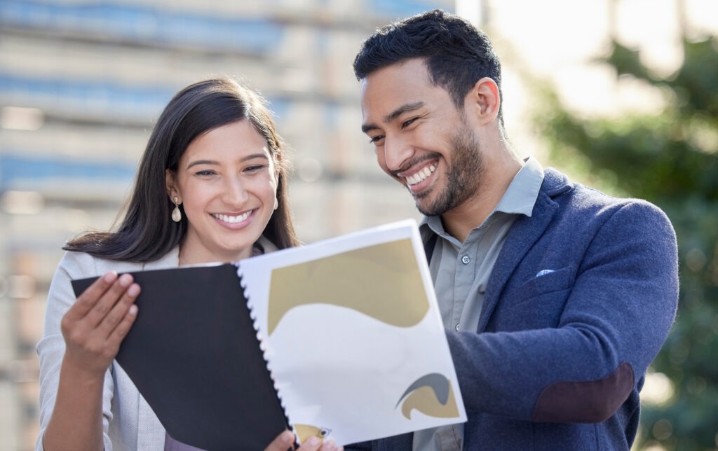 Two smiling individuals, a man and a woman, looking at a document together outdoors, discussing potential commercial loans.