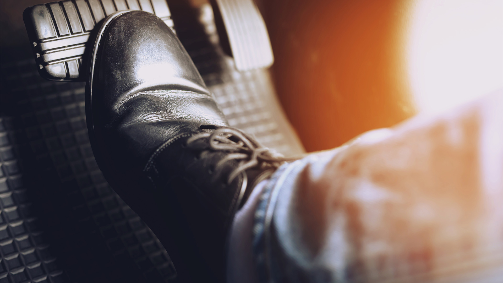 Close-up of a person's foot pressing a car's accelerator pedal. The person is wearing a black leather shoe and light blue jeans. The background features a warm, glowing light effect, reminiscent of the drive towards ambitious goals like those achieved with Business Loans tailored for success.